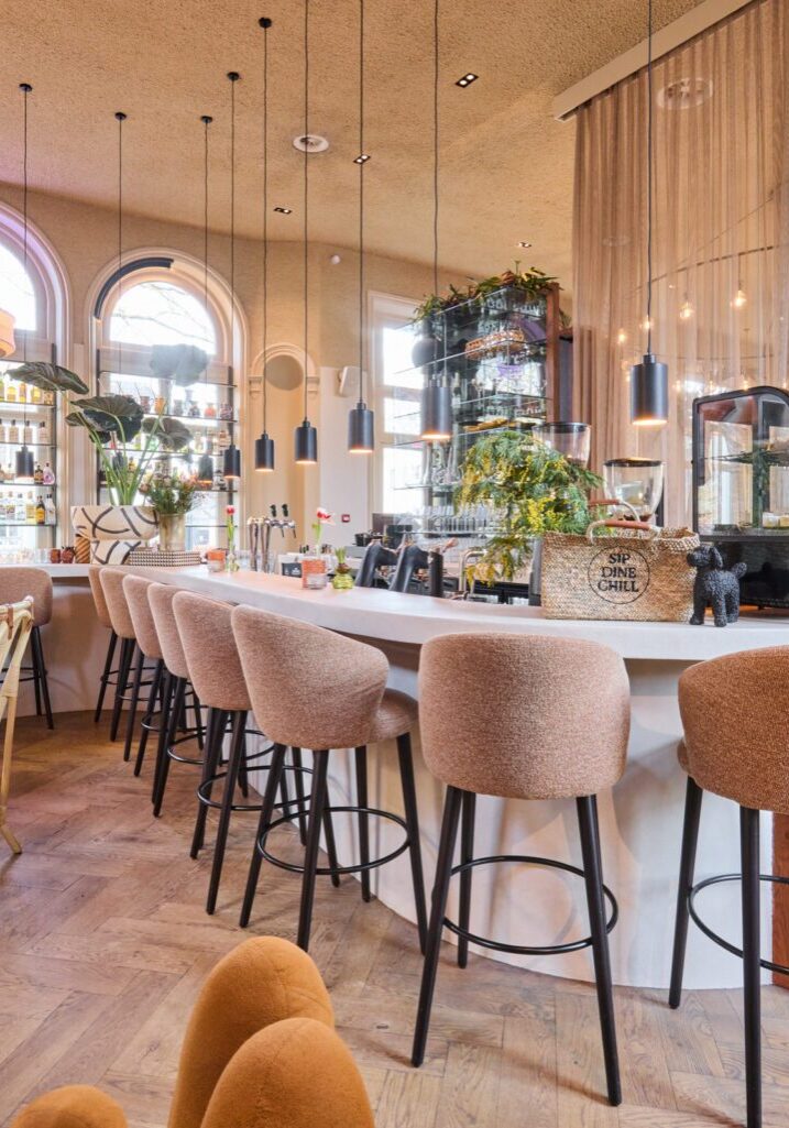 Vooraanzicht van de bar met microcement blad en houten zijkant, gestoffeerde barkrukken en stalen vitrineframes op de achtergrond